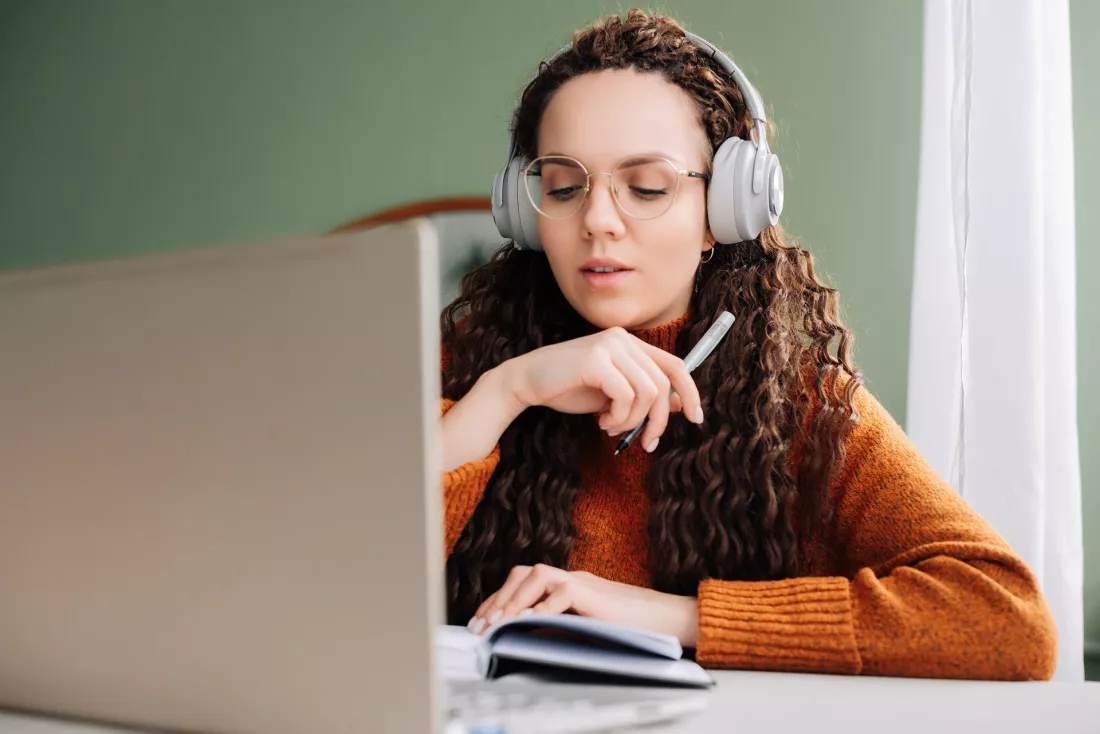 Studentka ze sluchawkami uczy sie online przy laptopie i robi notatki