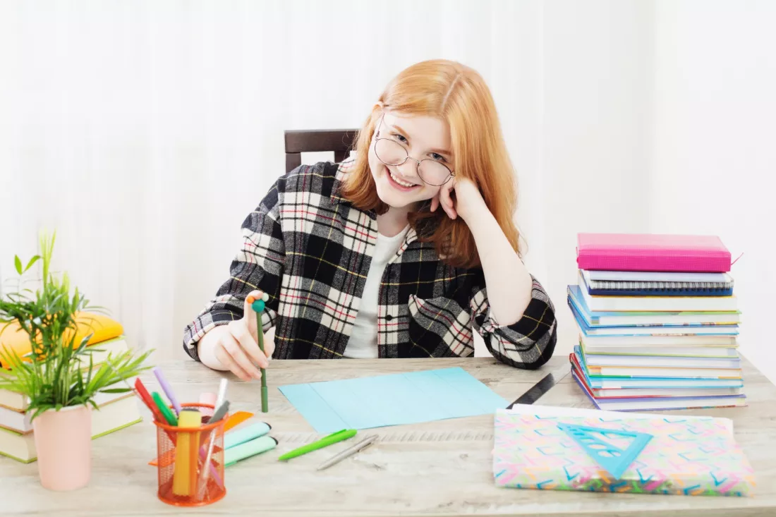 Usmiechnieta studentka w okularach uczy sie przy biurku z ksiazkami i kolorowymi notatkami