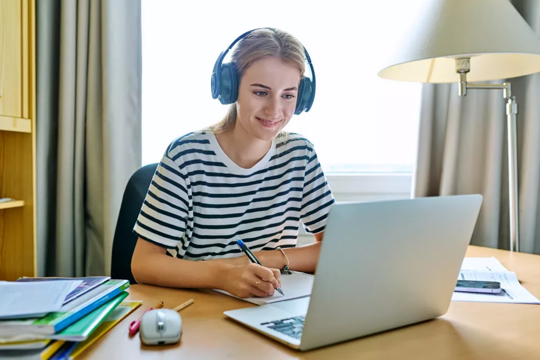 Mloda kobieta w sluchawkach uczy sie przy laptopie i robi notatki w zeszycie