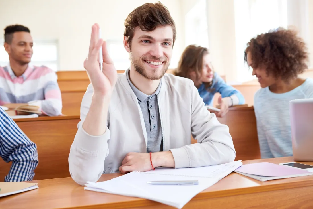 Uśmiechnięty student podnosi rękę, aby zadać pytanie podczas zajęć w sali wykładowej.