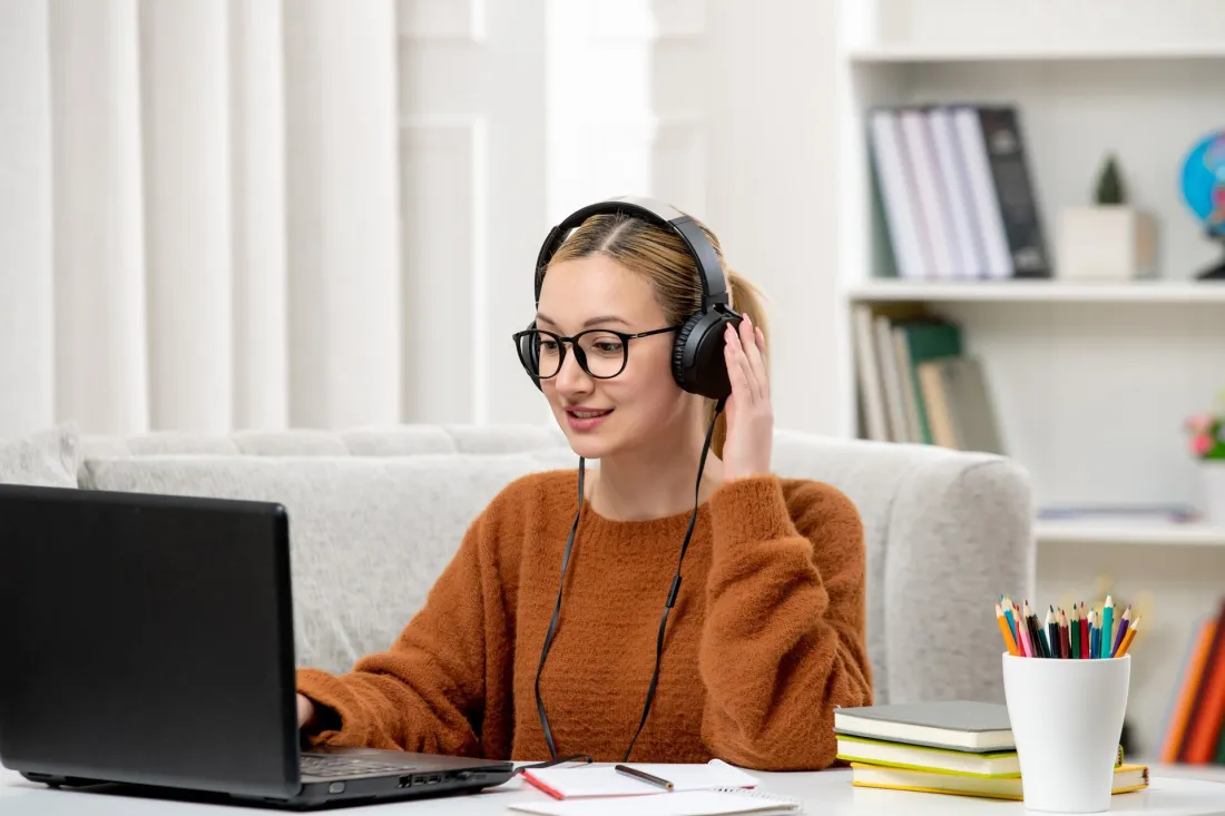 Studentka w okularach i słuchawkach uczy się przy laptopie w domowym pokoju, obok leżą notatki i kubek z kredkami.