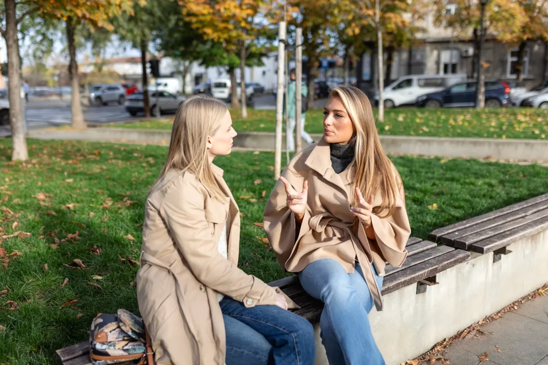 Dwie młode kobiety rozmawiają na ławce w parku jesienią, jedna opowiada, druga słucha